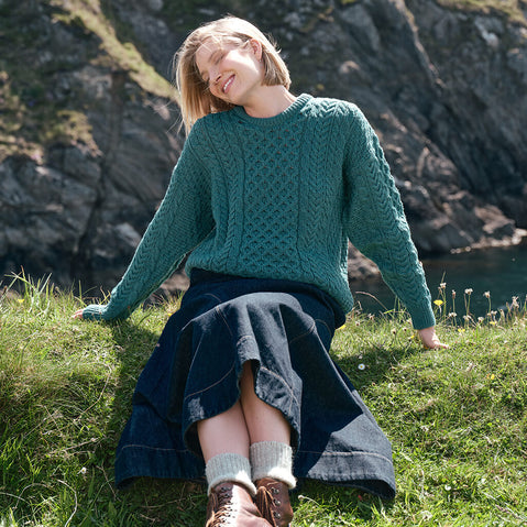 Women's Traditional Crew Neck Aran Sweater in Teal, featuring authentic Irish cable knit stitching and timeless craftsmanship, styled outdoors along the rugged Irish coastline.