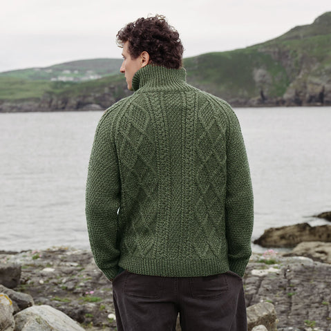 Back view of the Men's Hand Knit Aran Zipper Cardigan in green, crafted with authentic Irish Aran cable knit patterns, a full-zip fastening, and a high ribbed collar, showcasing traditional Irish craftsmanship against a scenic coastal landscape.