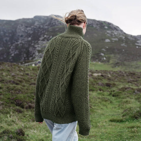 Back view of the Women's Hand Knit Aran Zipper Cardigan in green, crafted with authentic Irish Aran cable knit patterns, a full-zip fastening, and a high ribbed collar, showcasing traditional Irish craftsmanship against a scenic countryside backdrop.