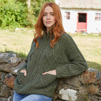 Woman wearing a green Women's Hand Knit Aran Zipper Cardigan sitting on a stone wall outdoors.