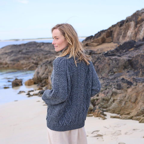 A woman with long, light brown hair stands on a sandy beach facing away from the camera. She wears a knitted, blue aran sweater and beige pants. In the background, rocky formations are visible beside the water. The sky is clear and adds to the serene coastal atmosphere.