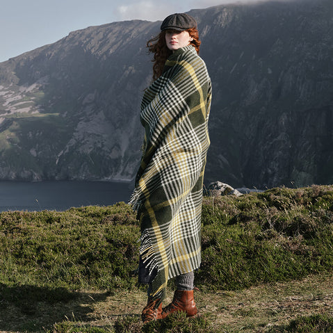 Image featuring the Foxford Michael Collins Wool Throw, crafted from luxurious wool with a timeless green, cream, and gold check pattern and fringed finish, wrapped around a person against a rugged Irish coastal landscape, showcasing heritage-inspired design and exceptional craftsmanship.