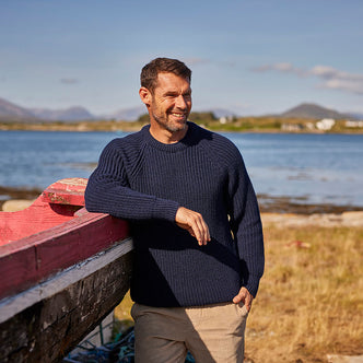 A man stands leaning on a boat by the coast wearing a traditional Irish fisherman sweater.