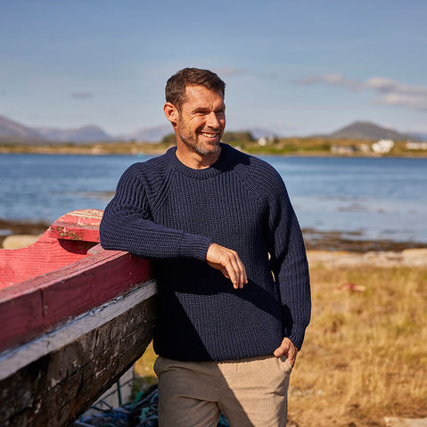 A man stands leaning on a boat by the coast wearing a traditional Irish fisherman sweater.