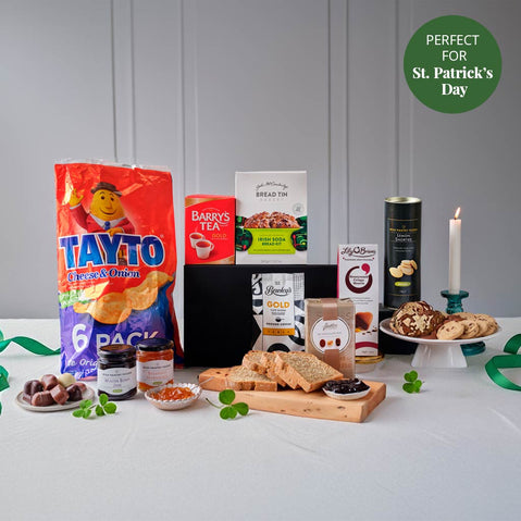 St. Patrick’s Day hamper featuring Irish treats including Tayto cheese and onion crisps, Barry’s Tea, soda bread mix, cookies, chocolates, jam, and gourmet snacks arranged on a table with shamrock decorations and a lit candle.