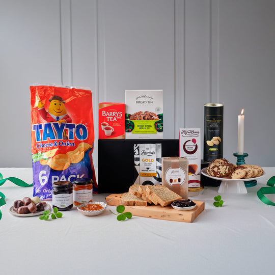 A curated gift basket arrangement on a white tablecloth with a gray paneled wall background. Visible items include a large bag of Tayto chips, a can of Barry's Tea, a loaf of bread, jars of spreads, cookies on a plate, and a black gift box, styled with green ribbon and candles.