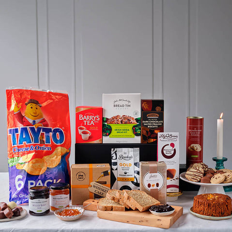 From Ireland with Love Food Gift Basket. A variety of food items displayed on a table. The image features a large bag of Tayto crisps, boxes of Barry's Tea and bread mix, jars of spreads, and various baked goods including biscuits and cakes. A lit candle is present, adding to the cozy atmosphere.