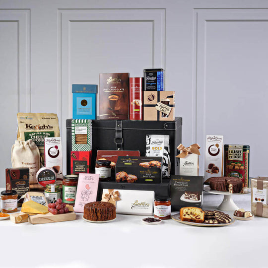 Irish Celebration Gourmet Gift Basket. A variety of gourmet food items arranged around a black gift box. The display includes chocolates, cookies, cheese, and jars of spreads. Some items are in boxes with elegant packaging, while others are placed openly, showcasing their contents. The background is neutral, highlighting the colorful assortment of treats.