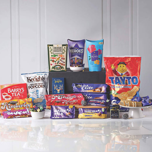Nostalgic Irish Favorites Gift Basket. A variety of snacks and Irish treats arranged on a white surface. In the foreground, there are packaged items including Barry's Tea, Kimberley biscuits, Tayto crisps, and Dairy Milk chocolate bars. Also featured are popcorn bags and assorted chocolates in colorful wrappers. The background is plain to highlight the products.