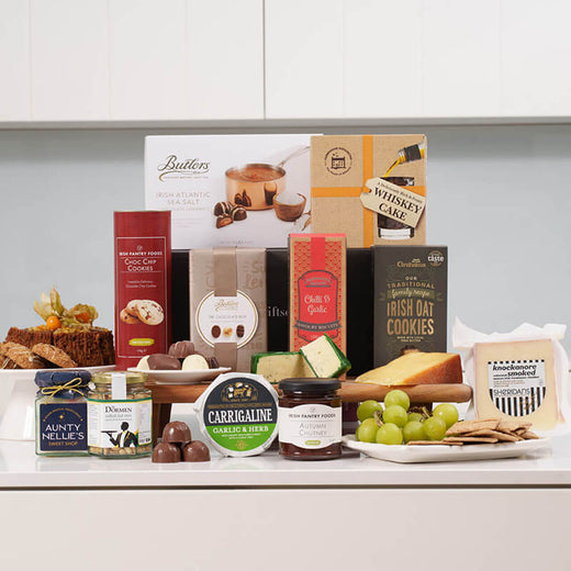 Irish Sweet & Savory Selection. An assortment of gourmet Irish snacks and treats is displayed on a white surface. Items include various chocolates, cakes, biscuits, cheese, crackers, and grapes. The arrangement features branded packaging prominently.