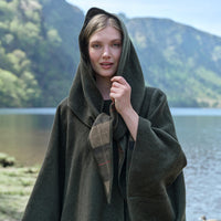 Woman wearing a green hooded cloak with a tartan pocket square, standing by a lake with mountains in the background.