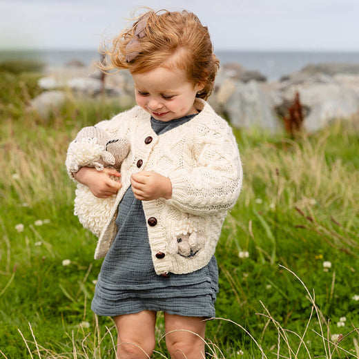 A young child stands in a grassy field, wearing a cream-colored kid's Aran cardigan with buttons and a blue dress. The child holds a small fluffy toy animal while looking down, surrounded by rocks and greenery in the background.