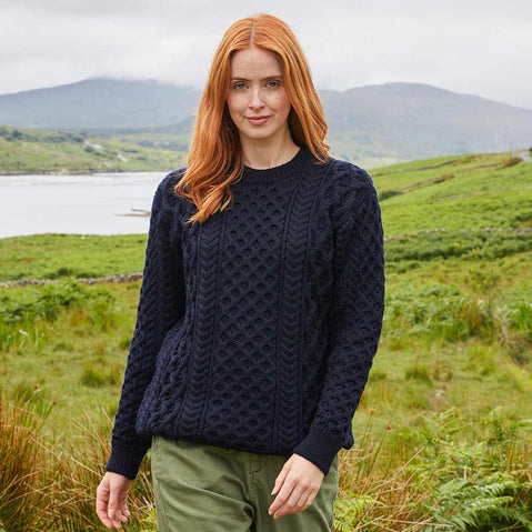 Woman wearing a navy blue Women's Crew Neck Irish Wool Classic Sweater in a scenic outdoor setting with green fields and mountains.