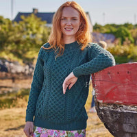 Woman wearing a green Women's Crew Neck Irish Wool Classic Sweater standing next to a red boat in a natural setting
