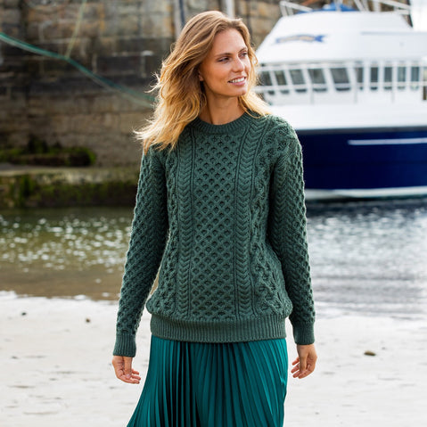 A woman standing by the water wearing a green Irish wool crew neck sweater.