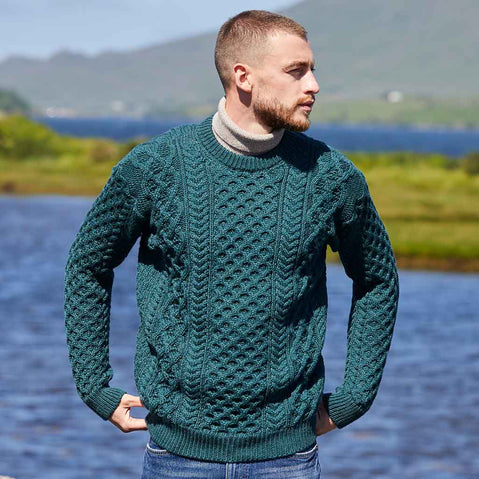 Man wearing a green Men's Crew Neck Irish Wool Classic Sweater standing by a body of water with mountains in the background