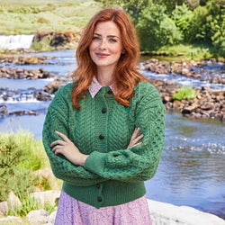 Image featuring The Cushendall Cropped Aran Cardigan in green, crafted with authentic Irish Aran cable knit patterns, a flattering cropped silhouette, and timeless button-front styling, showcasing traditional Irish craftsmanship against a picturesque riverside landscape.