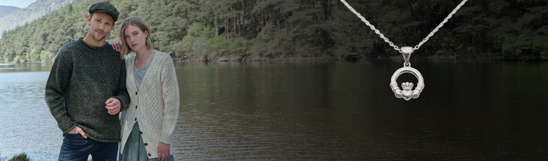 Couple wearing a patterned cardigan and fisherman sweater with a necklace featuring a circular pendant with a crown design, standing by a body of water with trees in the background.