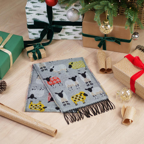 Grey fringed scarf with colorful sheep pattern displayed among wrapped Christmas presents, pine cones, and holiday decorations