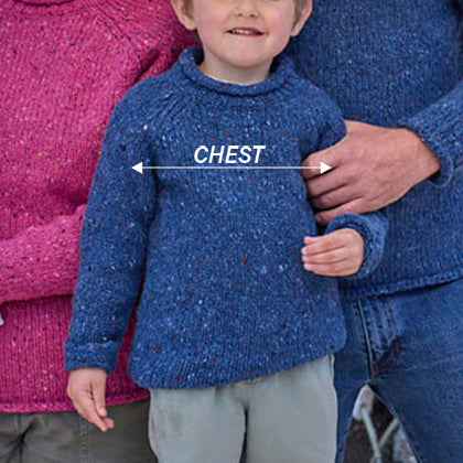 A young child wearing a blue knitted aran sweater stands beside an adult, who is also in a blue sweater. The child has a cheerful expression and is holding one arm slightly out. A label points to the child's chest, indicating the measurement area. In the background, there are blurred figures wearing pink and brown sweaters.