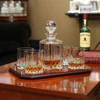 Irish Home Decor - A polished wooden tray holds a crystal decanter topped with a faceted stopper, surrounded by four short crystal glasses. The glasses contain amber liquid. In the background, a bottle of Jameson whiskey is visible on a shelf, alongside a blurred setting that suggests a cozy, well-furnished room.