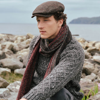Peaky Blinders Wool Cap in Brown Red, featuring a classic heritage tweed design and timeless Irish craftsmanship, styled alongside traditional Irish knitwear and a luxury alpaca scarf against the rugged Irish coastline.