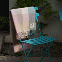A graqy/biege plaid blanket drapes over a turquoise metal chair, set against a backdrop of green foliage and a textured surface.