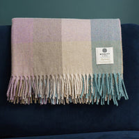A folded, checked blanket in soft pastel colors rests on a dark blue surface. The blanket features fringed edges. The background is a muted green.