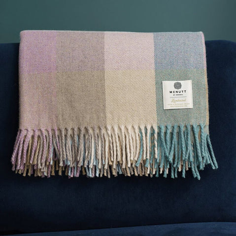 A folded, checked blanket in soft pastel colors rests on a dark blue surface. The blanket features fringed edges. The background is a muted green.