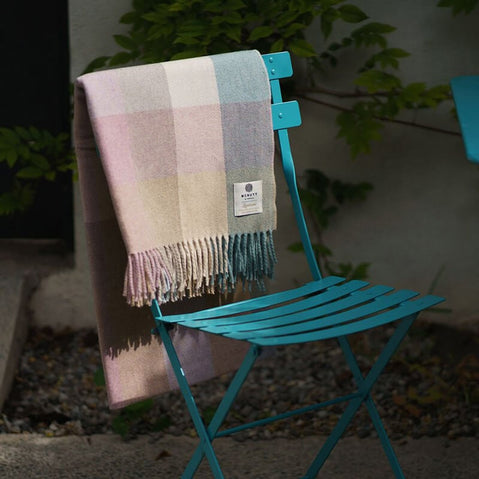 A graqy/biege plaid blanket drapes over a turquoise metal chair, set against a backdrop of green foliage and a textured surface.