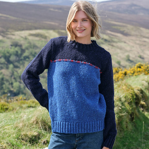 Woman wearing a blue and navy sweater standing in a scenic outdoor setting with hills and flowers.