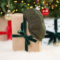 Olive green Irish tweed flat cap with herringbone pattern resting on kraft paper gift box tied with dark green velvet ribbon, surrounded by Christmas decorations and bokeh lights