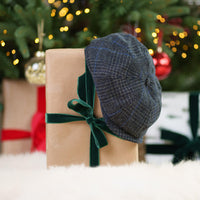 Navy blue Irish tweed flat cap with herringbone pattern displayed on kraft paper gift box with dark green velvet bow, festive Christmas setting with ornaments and twinkling lights in background