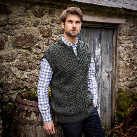 A man stands in front of a stone wall, wearing a green Aran vest over a blue and white checkered shirt. He has short brown hair and appears smiling & relaxed. He leans against an empty wooden barrel beside the stone structure.