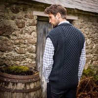 A man stands with his back to the camera wearing a dark knitted Aran vest over a blue and white checkered shirt. He has short brown hair and appears relaxed, with his hands placed in front of him. In the background, there is an empty wooden barrel beside a stone structure.