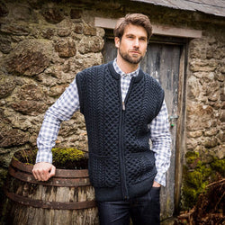 A man stands in front of a stone wall, wearing a dark knitted Aran vest over a blue and white checkered shirt. He has short brown hair and appears smiling & relaxed. There is an empty wooden barrel beside the stone structure.