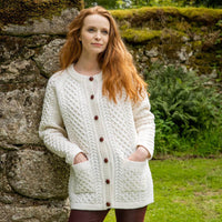 A woman standing by a stone wall wearing a cream The Irish Store Women's Aran Button Cardigan with cable and basket stitch details.