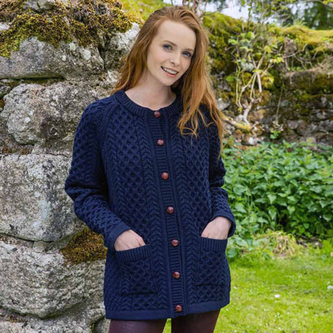 Woman wearing a navy blue The Irish Store Women's Aran Button Cardigan with red buttons against a stone wall and greenery background.