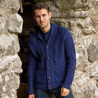 Man wearing a navy The Irish Store Men's Classic Aran Cardigan standing against a stone wall.