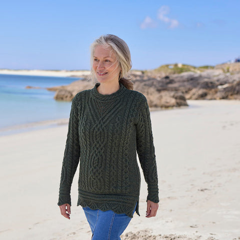 Woman wearing an army green Women's Signature Aran Tunic Sweater walking along a beach with a clear blue sky. 