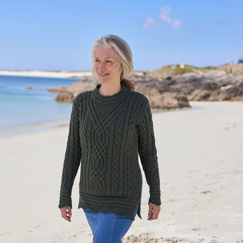 Woman wearing an army green Women's Signature Aran Tunic Sweater walking along a beach with a clear blue sky. 
