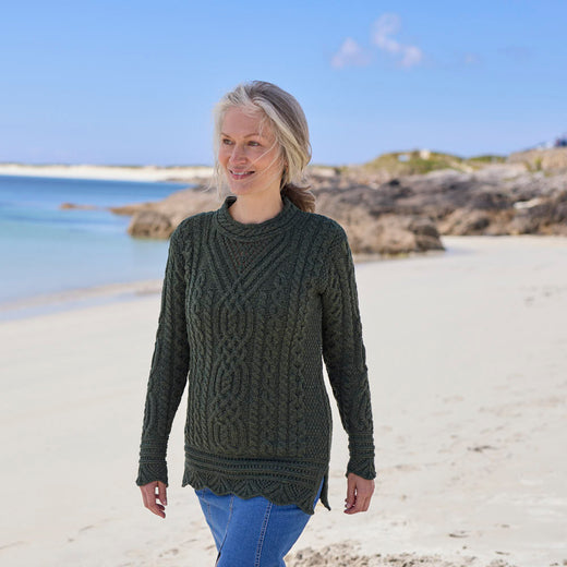 Woman wearing an army green Women's Signature Aran Tunic Sweater walking along a beach with a clear blue sky. 