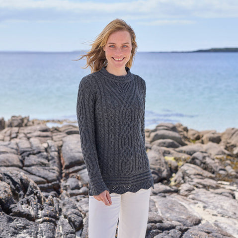 Woman wearing a charcoal gray Women's Signature Aran Tunic Sweater standing by a rocky shoreline with a clear blue sky. 