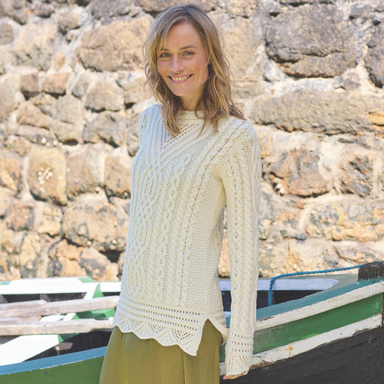 Woman wearing a cream Women's Signature Aran Tunic Sweater standing in front of a stone wall and boat.