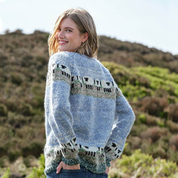 A person with blonde hair wearing a blue Women's Mohair Sheep Cardigan featuring sheep patterns around the upper part and near the hem, standing outdoors with a scenic background of hills and greenery.