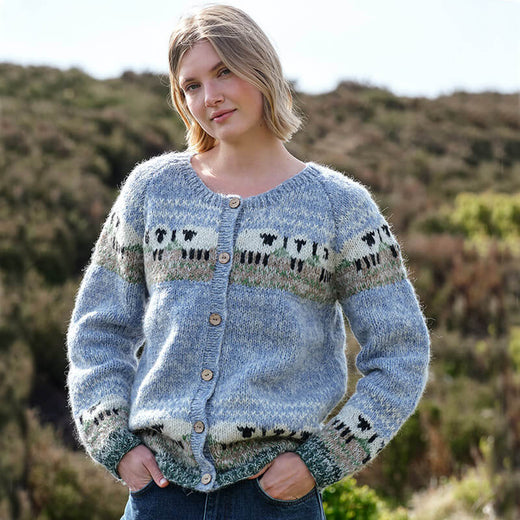 A person wearing a light blue Women's Mohair Sheep Cardigan with black, white, and green sheep and geometric patterns across the chest and sleeves. The cardigan features wooden buttons and a soft, fuzzy mohair texture, set against an outdoor backdrop of greenery and hills.