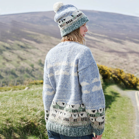 Person wearing a patterned sheep sweater and matching hat standing on a grassy hillside with a scenic background.