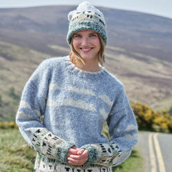 Woman wearing a patterned sheep sweater and matching hat, standing in a scenic outdoor setting with mountains and a road.