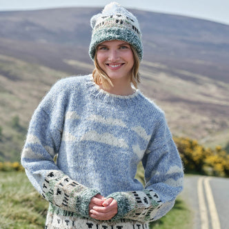 Woman wearing a patterned sheep sweater and matching hat, standing in a scenic outdoor setting with mountains and a road.