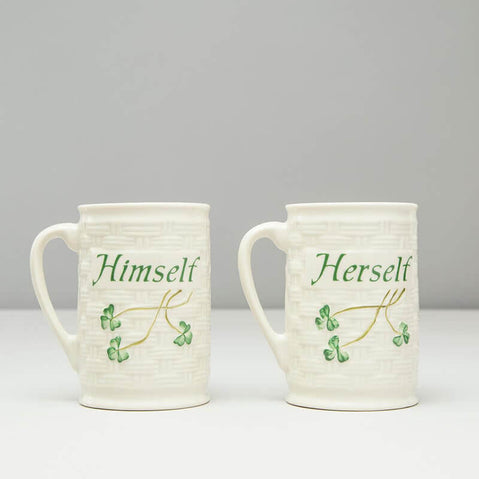 Two Belleek china mugs are presented side by side, one labeled Himself and the other Herself, both featuring green lettering and a subtle leafy design. The background is plain and neutral in color.